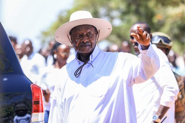 President Yoweri Museveni of Uganda, wearing a light-colored hat and white long-sleeved shirt, raises his hand in front of crowd of people, whose images are blurred.