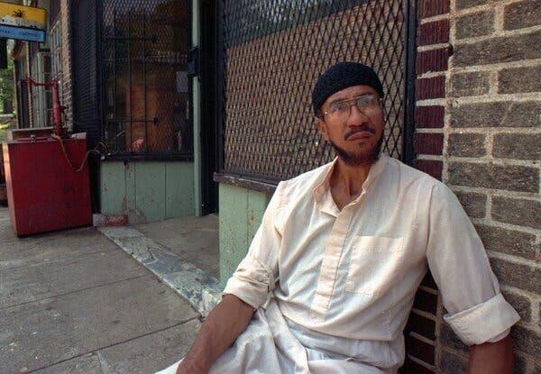 A man in a white shirt leans against a brick wall.