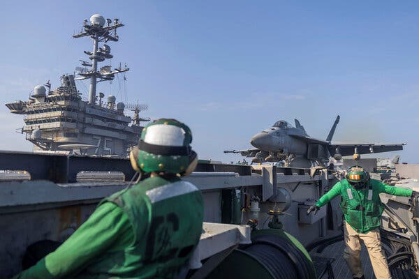 Two people wearing green helmets and flight suits with ear protection stand near a fighter jet aboard an aircraft carrier.