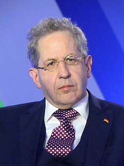 close-up of Hans-Georg Maaßen wearing a white shirt, purple-and-white checkered necktie, and dark jacket with a lapel pin of a German flag, looking slightly left of camera