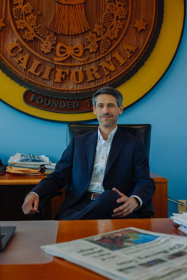 Matt Mahon, the mayor of San Jose, Calif., sits in an office chair at his desk, with a big wooden city seal behind him on the wall. 