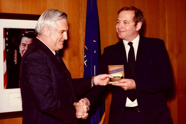 Mr. Johnson, who has brown hair, shakes hands with a gray-haired man, who has handed him a medal in a small box. A photo of President Ronald Reagan is on a paneled wall behind them.
