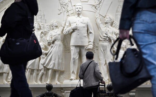 People in a subway station look at a relief of Joseph Stalin that depicts figures on either side of him, holding up flowers.