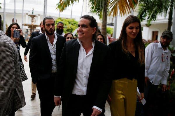 Alex Saab, in a dark suit and white shirt, walks among a group of people as they past palm trees and a white-domed building.