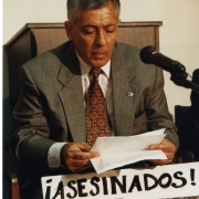 José Basulto in a color photo, reading in a photo in front of a microphone and sign that reads, "Asesinados!"