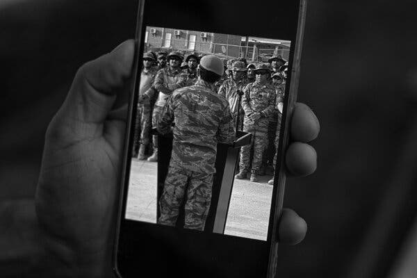 A hand holding a cellphone, which contains an image of a man in fatigues addressing others.