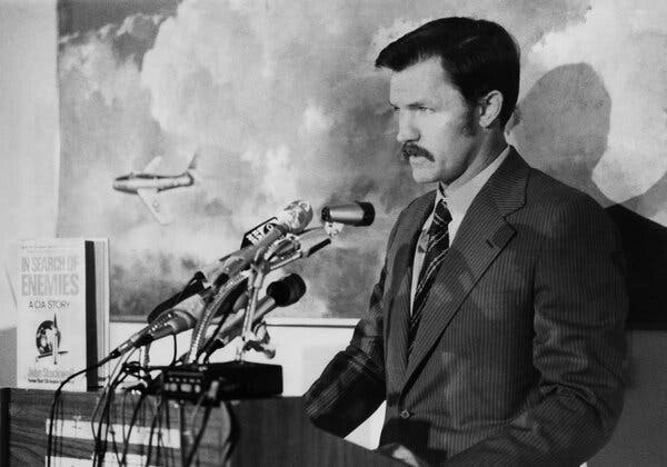 A black-and-white photo of John Stockwell with short, dark hair, long sideburns and a mustache, standing at a podium in front of a stand of microphones next to his book.