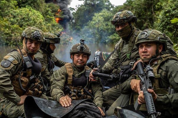 A group of people in military gear sit in a boat. In the background, a fire emits thick black smoke over the green jungle and river.