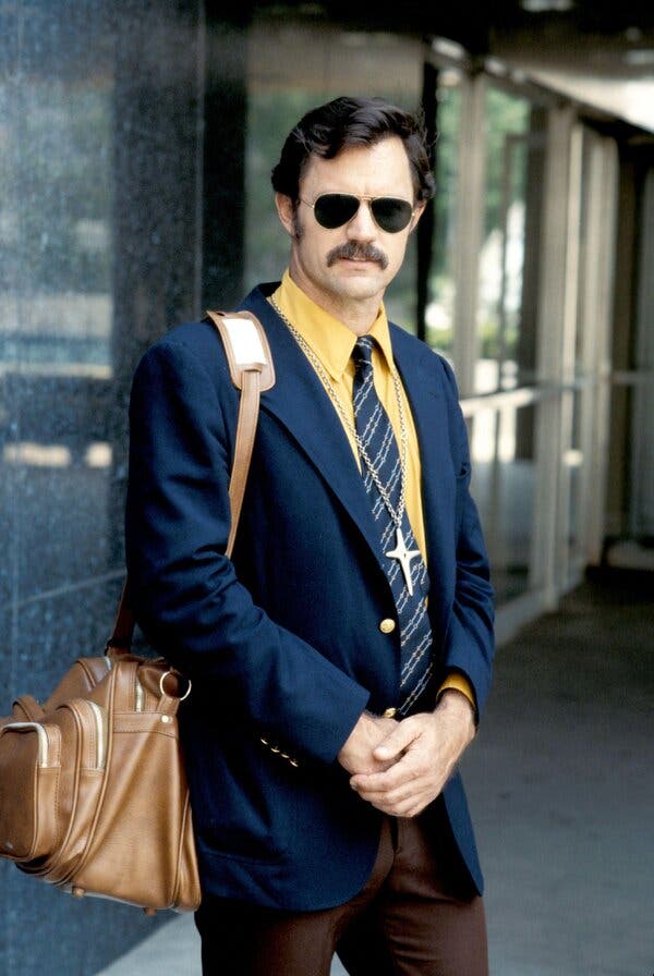 John Stockwell, with short, dark hair and a mustache, stands outside a marble building, wearing dark aviator glasses, a yellow shirt, striped tie and blue blazer, carrying a brown messenger bag.