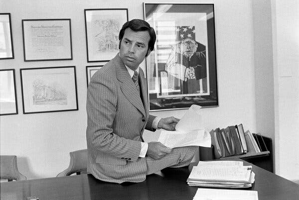 A black and white photo of Mr. Pottinger wearing a light-colored pinstriped suit and sitting on an office desk while holding papers.