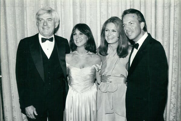 A black and white photo of two couples standing shoulder to shoulder against a curtain backdrop. The men wear black tuxedos and the women wear gowns.