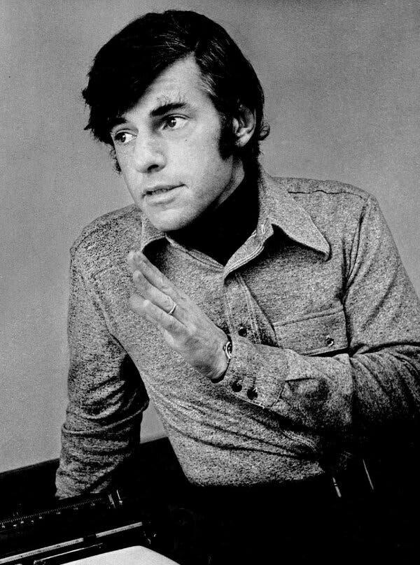 A black-and-white portrait of H. Bruce Franklin, a man with long hair wearing a button-down shirt while gesturing with his left hand and looking away from the camera.