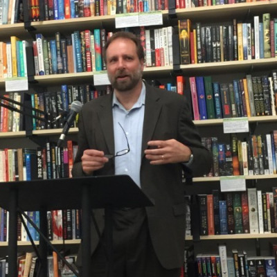 A candid photo of Stephen standing at a podium speaking into a microphone with a tall and colorful bookshelf behind him