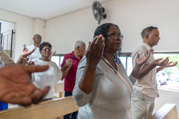 A group of churchgoers stand with open hands raised. Roseline Rabouine wears glasses and a light grey top. Her mouth is slightly open.