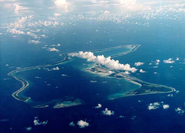 An aerial view of Chagos Archipelago, a narrow, ring-shaped island chain in a deep blue ocean, encircling a turquoise lagoon. White clouds dot the sky.