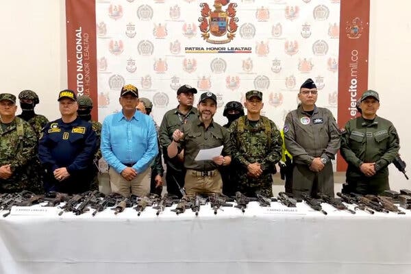Colombia’s president, President Abelardo De La Espriella, stands before a table covered with guns, gesturing with one hand as he holds notes in his other hand. People, some in military uniforms, stand around him.