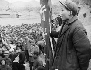 Mao Zedong stands on a raised platform, speaking to a large crowd of followers gathered outdoors. Mao is in profile, holding a vertical banner with Chinese characters.