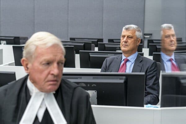 In a setting with rows of black computer monitors, a man in a black robe is in the foreground. A man in a dark suit and red tie sits behind him, looking ahead.