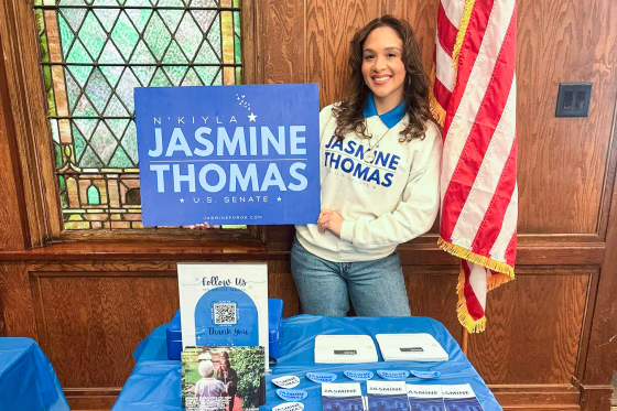 N’Kiyla Jasmine Thomas stands in front of a campaign table while holding a blue sign that reads "N’Kiyla Jasmine Thomas, U.S. Senate"